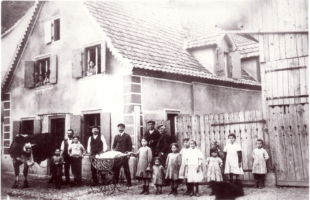 A large group of adults and children standing in front of a house, with a cow and a wheelbarrow