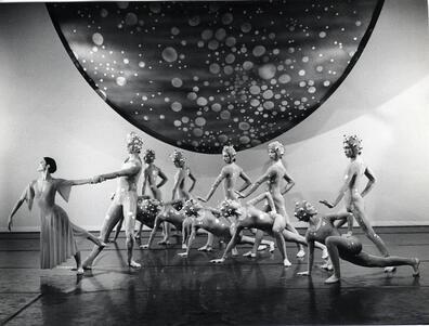 Twelve dancers onstage. Eleven wear leotards and hair caps with metallic balls attached to them, one wears a dress and leans away from the others.