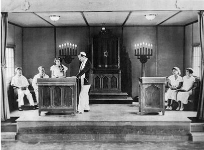 A picture of a bimah, with two men and two women sitting on either side, and a man in a kippah and prayer shawl standing by a podium, and a young woman with flowers pinned to her dress speaking at the podium