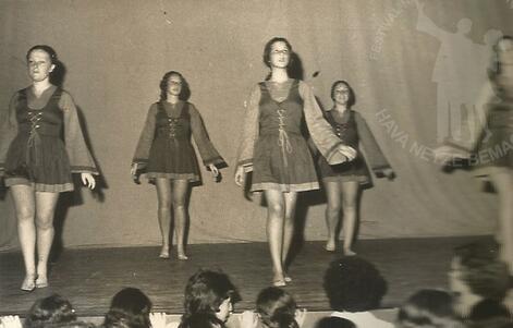 A group of five women wearing folk dancing costumes, performing barefoot on stage