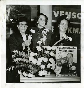 Ida Weis Friend and two other woman with a vase full of flowers, beside a sign reading "Wear a white carnation for STEVENSON. VOTE and show your colors!"