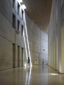 A curved hallway of simple grey stone, with very high ceilings and skylights