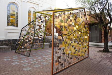 An asymetrical metal sukkah set up in a courtyard. One side is fine yellow mesh, while the others are a looser grid with photographs of eyes and printed writing filling some of the gaps.