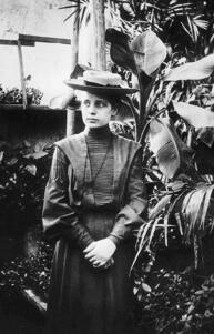 Lise Meitner standing with her hands folded in a greenhouse beside a large tropical plant