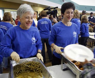 Lynn and Stacy Schusterman serving food, wearing blue volunteer shirts with a large group of other volunteers in the background