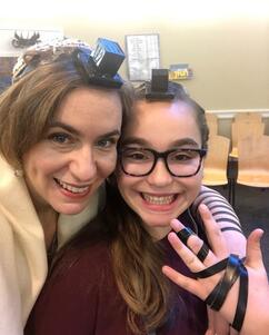 Ma'ayan Rosenbaum, holding up a hand to show the straps of her tefillin, with her mother standing beside her with an arm around her shoulders, wearing tefillin, tallit, and kippah