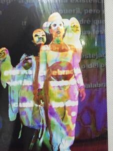 Yemile Misrahi on stage, wearing a dance costume and a plain white domino mask, standing in front of another dancer wearing and holding up more dramatic masks, with colorful lights and text projected on them