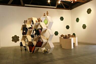 A gallery exhibit with a set of octagonal shelves containing exhibit items, and a collection of clay pottery on a set of displays