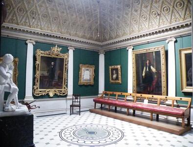 A room with a tiled floor and arched ceiling, with a long bench for visitors, paintings on the walls, and a small white sculpture in the foreground