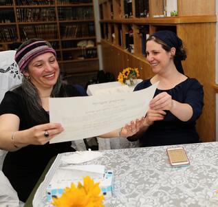 Two women seated at a table in a library, holding up a large piece of paper