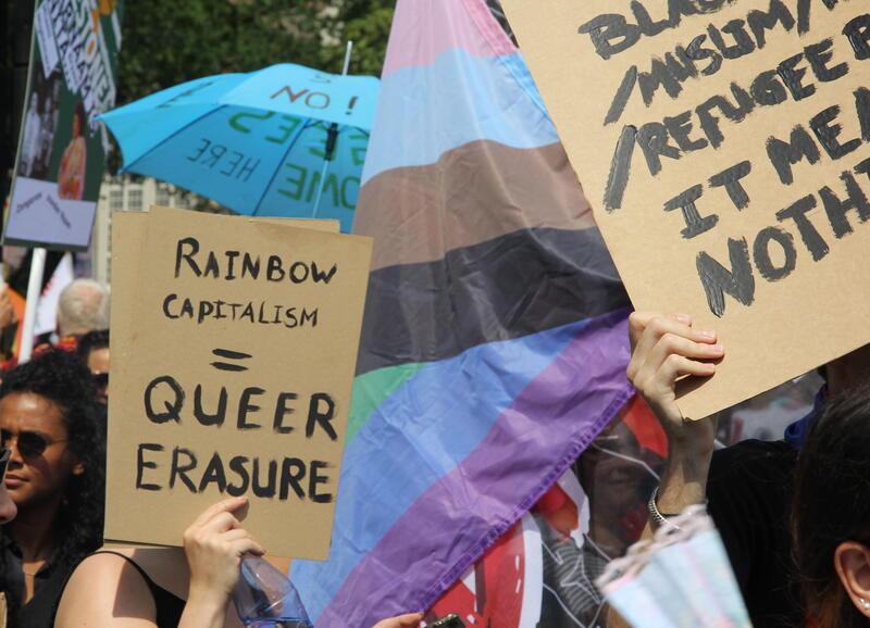 Image of a march at Amsterdam pride: figure holding up sign that reads: "Rainbow Capitalism = Queer Erasure"