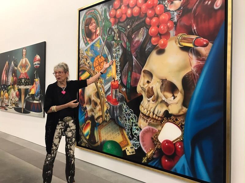 Audrey Flack wearing acid wash jeans and a black top standing in front of a large painting with many items including a skull, tarot, lipstick, candle