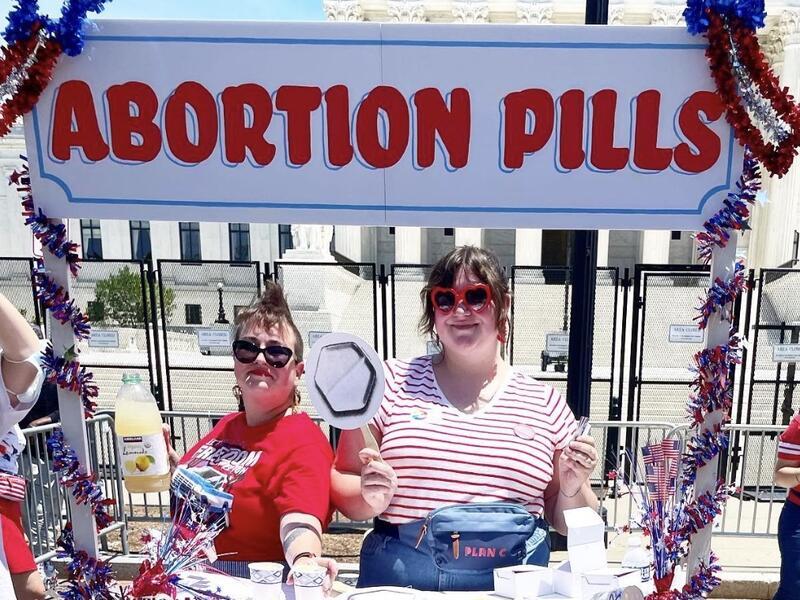Two women stand at a booth with a sign that says "Abortion Pills"