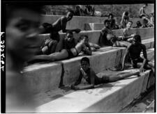 Black-and-white photograph of Black children lounging on concrete steps in bathing suits, looking at photographer, blurred figure on edge of photo