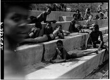 Black-and-white photograph of Black children lounging on concrete steps in bathing suits, looking at photographer, blurred figure on edge of photo