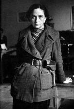 Mika Feldman de Etchebéhère leaning back against a desk, wearing a belted coat with a holster