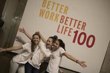 Three young women posing in front of a sign that reads Better Work Better Life 100 in yellow, orange, and red text on a white background