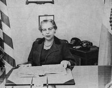 Anna Moscowitz Kross sitting at a desk with a file open in front of her. In the background are two telephones, a diploma on the wall, and the flags of the United States and New York City
