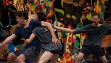 Three dancers in street clothes pulling at each other on a stage, in front of a tattered, colorful patchwork background