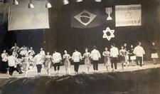 A line of dancers, men and women alternating, performing onstage with musicians. In the background are an Israeli flag, a Brazilian flag, and a banner with a menorah and Star of David