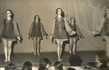 A group of five women wearing folk dancing costumes, performing barefoot on stage