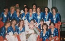 A group of dancers posing backstage, wearing blue, red, and white costumes, with one woman in the center wearing a white dress and holding a bouquet of red roses