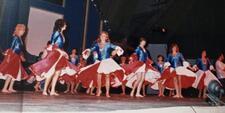 A group of dancers onstage, wearing costumes with blue tops and wide white and red skirts