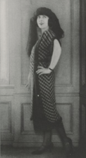 Sally Milgrim posing in a calf-length dress with a plaid pattern, black mary jane shoes, and a turban-style hat with fabric draped from it