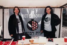 Two women in jackets and t-shirts at a booth. The poster behind them and their t-shirts both have a pomegranate logo and the words "Bet Debora" in Latin and Hebrew alphabets.