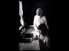 Portrait of an older woman with a white headscarf and a young girl, standing by a kitchen sink
