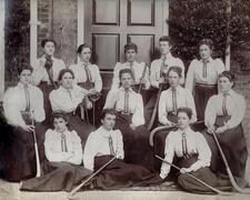 A group of young women in uniform dresses with field hockey sticks