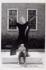 Janet Indick sitting on an outdoor stone plinth, on which sits an abstract statue of curving lines