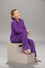 Judy Sheindlin smiling to camera while sitting on grey prop stairs in purple suit and heels in front of grey backdrop