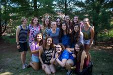 14 young women smiling for a group photo with some standing and some squatting, outdoors surrounded by trees