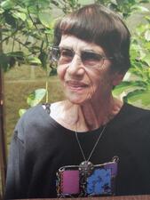 Color portrait of Judith Shuval against background of trees and plants and brick wall