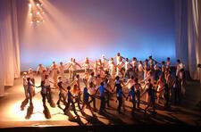 Wide shot of a large group of dancers in white t-shirts, holding hands in conentric circles around one dancer lifting a hand to the sky