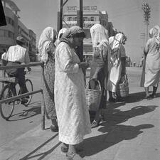 A half-dozen women stand on a sidewalk carrying baskets, waiting.