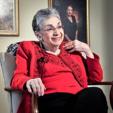 Meyera Oberndorf smiling to someone off camera and seated in front of portraits wearing a red top and black pants 