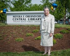 Meyera Oberndorf stands in front of balloon decorated outdoor sign for a library reading: Meyera E Oberndorf Central Library Virgina Beach Public Library