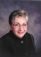 Portrait of Meyera Oberndorf smiling wearing glasses and a black jacket against a neutral background