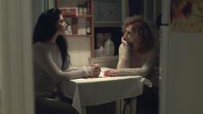 Two women sit at a small table in a dimly lit kitchen looking at one another