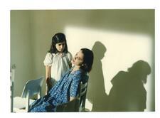 A child in a white dress stares at a women in a blue dress slouched in a chair looking out of frame 