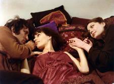 Three people lying in bed. A man, left, and woman, center, looking into each others eyes while a woman, right, holds the hand of the other 