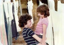 A boy with curly hair in a blue striped shirt lifting up a girl with shoulder length hair in a pink shirt. They are between clothes hang drying on lines.