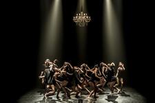 A large group of dancers in black leotards, and some in black jackets, dancing on a bare stage beneath a chandelier