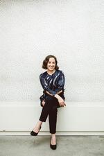 Niki Russ Federman smiling, seated with legs crossed on a white bench in front of a white textured wall