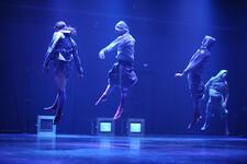 Four dancers in hooded sweatshirts leaping in the air, with the stage lit blue