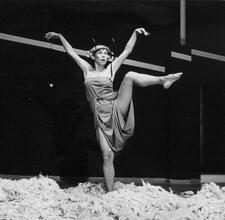 Ruth Eshel standing in a pile of loose wool, with one leg and both arms lifted high, wearing a thick iron circlet around her head.