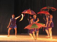 A group of dancers on stage, all wearing loose blue/purple velvet shorts and shirts. Four are holding colorful red, red/purple, and red/green umbrellas, one holds a horse hair whip