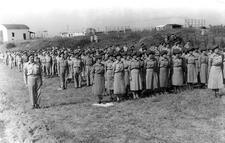 A large group of soldiers standing at attention in a rural area. At the front stand two lines of women soldiers in uniform dress and berets.